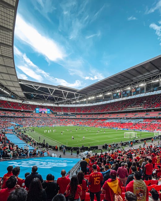 A panoramic view of Wembley Stadium filled with spectators watching a football match on a lush green pitch. The stadium is covered by a large, modern roofing structure, and the sky above is bright blu